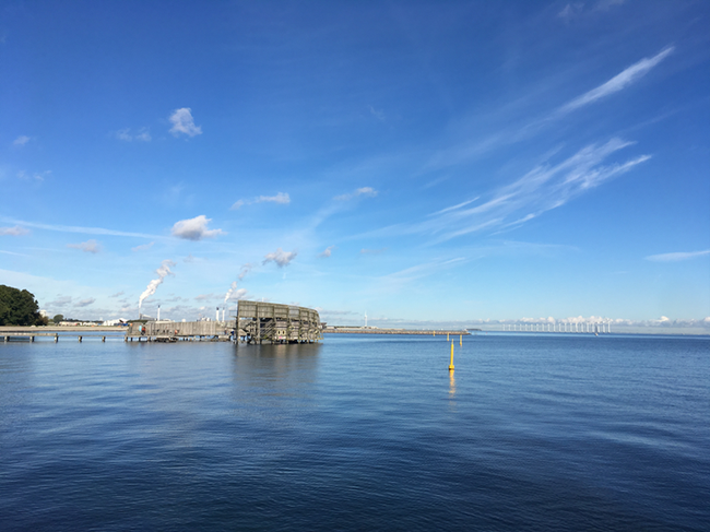 Figure b: The chimneys of the Danish powerplant “Amagerværket” in the background of Kastrup Sea Bath. Previously a coalfired powerplant from 1971, modified in 2020 to accommodate wood-pellets as main source of renewable energy, with gas and coal as backup. The powerplant supplies 650 MJ/s heat and 218 MW electricity to households in the Copenhagen area, supplemented by windmills.